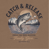 A brown t-shirt featuring a circular graphic print of a largemouth bass jumping out of water, with mountains, pine trees, birds, and a sun behind it. The text reads 'CATCH & RELEASE' and 'FREELY YOU HAVE RECEIVED, Freely Give.